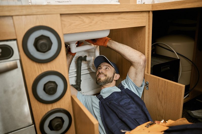 Cheerful plumber fixing sink pipe in kitchen